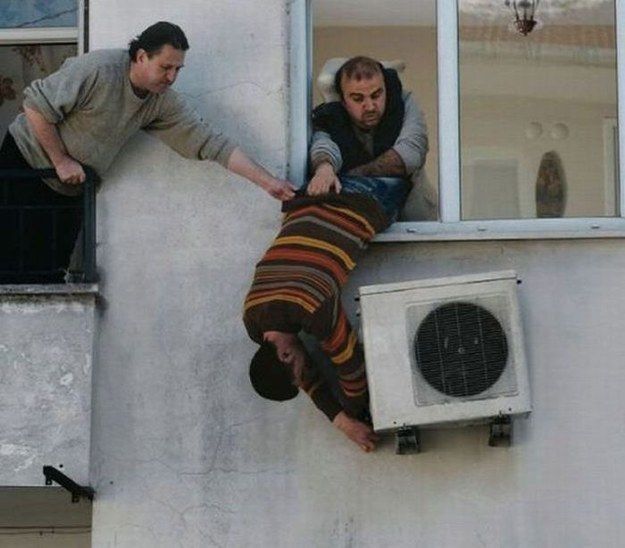 Three men hanging out of a window trying to fix an AC unit.