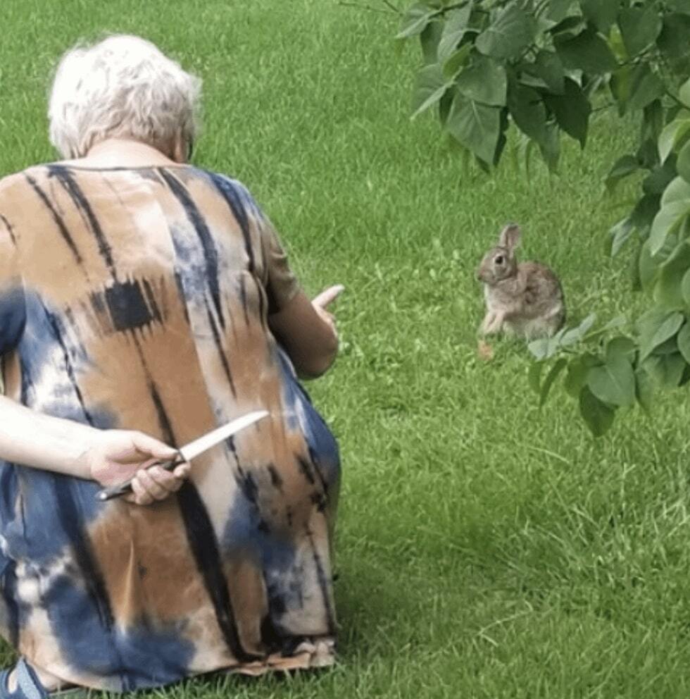 An old lady holding a knife behind her back while pointing at a rabbit