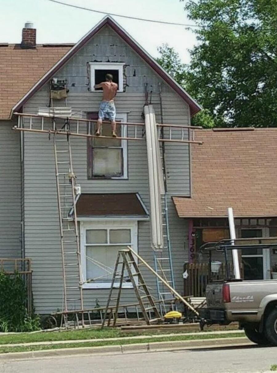 A guy unsafely standing on three different ladders to build a house
