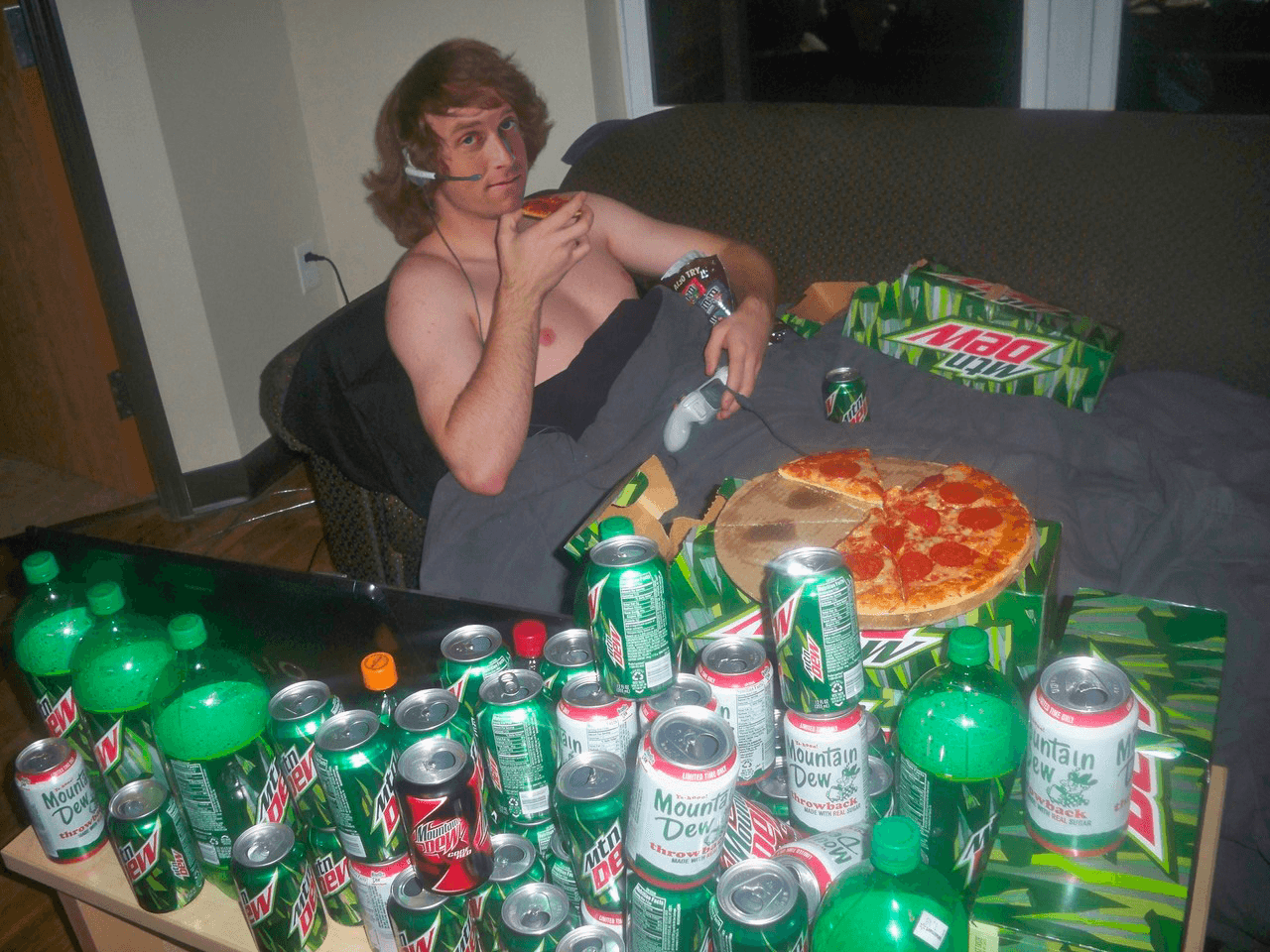 A shirtless man wearing a gaming headset eats pizza surrounded by Mountain Dew cans