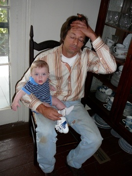 A tired man holding a baby while seated in a chair